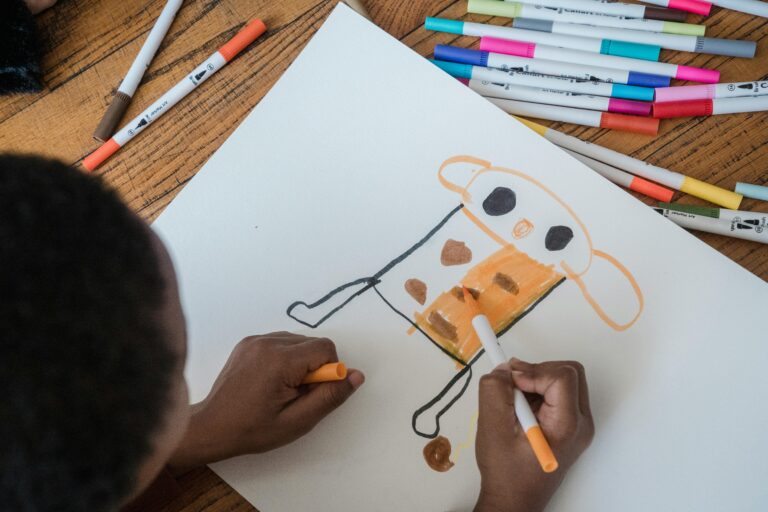 A child uses colored pens to draw and color a creative figure on white paper.